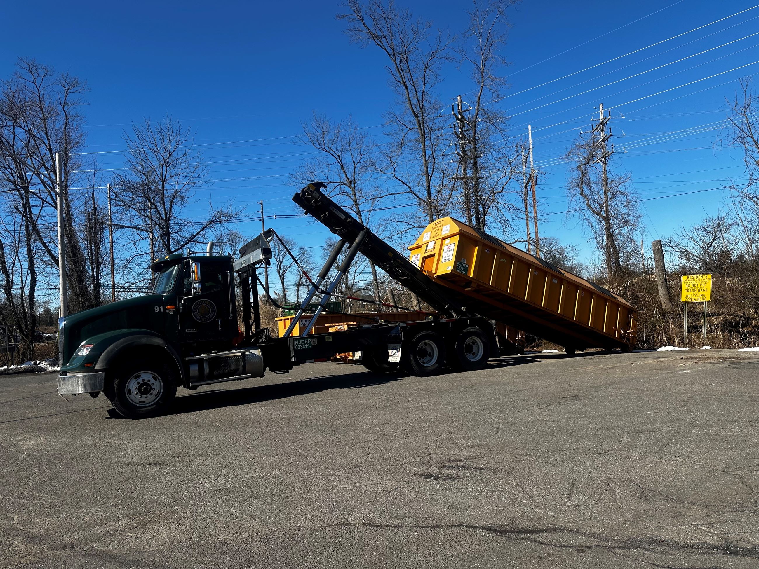 Green garbage truck with an extendable crane and brown dumpsters parked on a paved road beside utility poles.